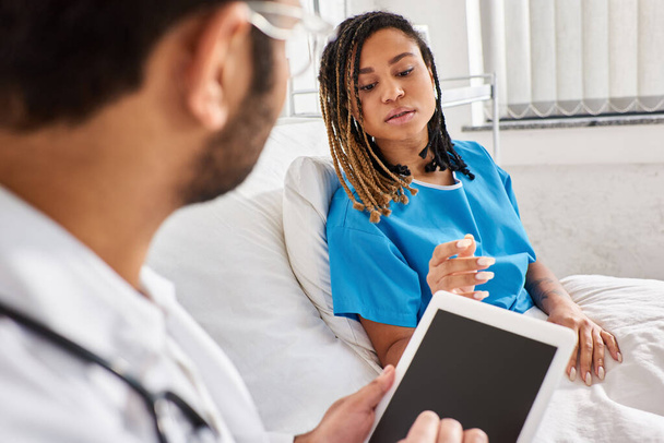 Afro-Amerikan hasta, hastane yatağında yatıp Hintli doktoruyla konuşuyor ve tablete not alıyor. - Fotoğraf, Görsel
