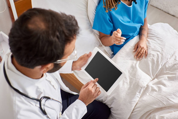paciente afroamericana sentada en la cama cerca de su médico indio tomando notas en la tableta, recortada - Foto, Imagen