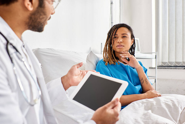 fókuszálni afro-amerikai nő kórházi ágyban nézi az indiai orvos jegyzetel tabletta - Fotó, kép