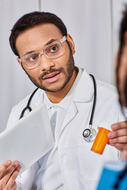 Fokus auf indischen Arzt in Brille zeigt Pillen seinem afrikanisch-amerikanischen Patienten mit Tablette - Foto, Bild