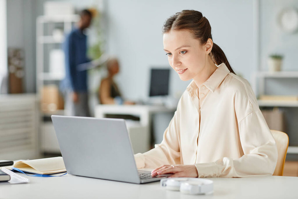 Портрет улыбающейся девушки, использующей ноутбук на рабочем месте в минимальном интерьере, копировальное пространство - Фото, изображение