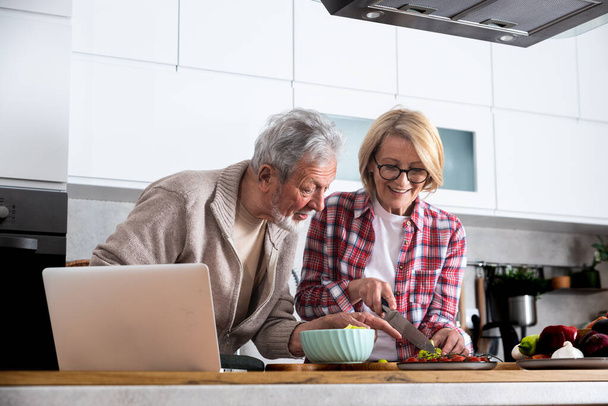 Idősebb pár vacsorát főz együtt a konyhában az arany házassági évfordulóra, receptet olvasnak az internetről a laptopon. Idősebb férfi és nő ételt készít a családnak, hogy megünnepeljék a szerelmet.. - Fotó, kép