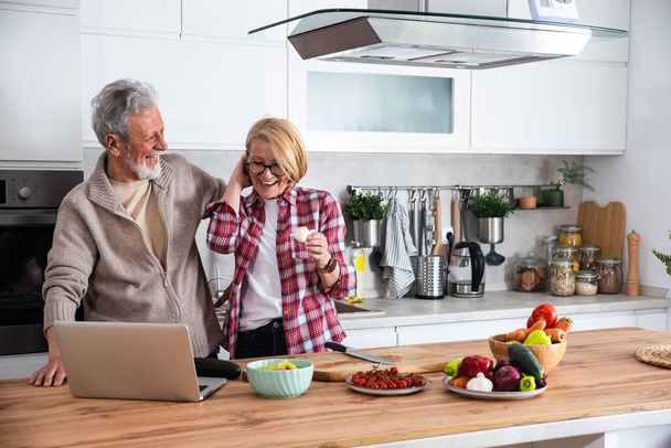 Seniorenpaar kocht gemeinsam in der Küche zum goldenen Hochzeitstag, liest Rezept aus dem Internet auf Laptop. Ältere Männer und Frauen, die Essen für die Familie kochen, laden sie ein, die Liebe zu feiern. - Foto, Bild