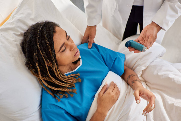 centrarse en la mujer afroamericana acostada en la cama mientras que el médico indio le enseña a usar inhalador - Foto, Imagen