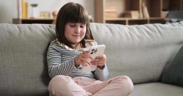 Cheerful girl sits on the living room couch, holding her phone with a bright smile. Her happy expression adds a sense of positivity to the room, creating a delightful scene. - Footage, Video