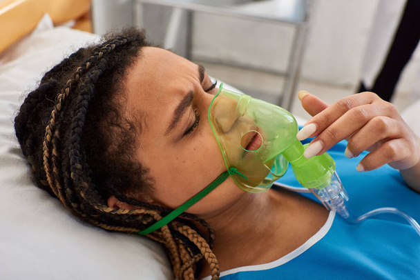 mujer afroamericana joven enferma acostada en la cama de hospital en su sala con máscara de oxígeno, atención médica - Foto, Imagen