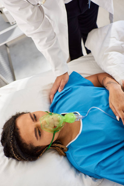 vista recortada de médico reconfortante mujer afroamericana enferma acostada en la cama de hospital con máscara de oxígeno - Foto, Imagen