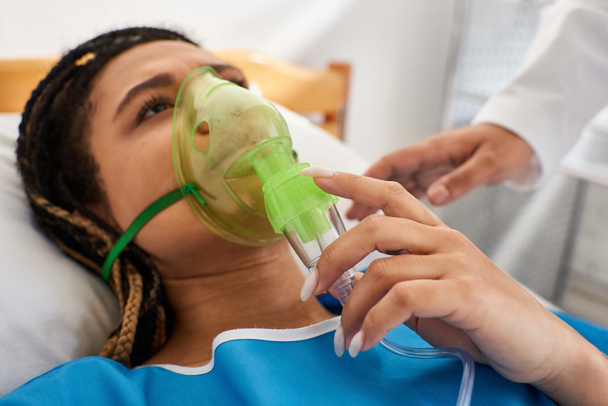 vista recortada de médico reconfortante mujer afroamericana enferma acostada en la cama de hospital con máscara de oxígeno - Foto, Imagen