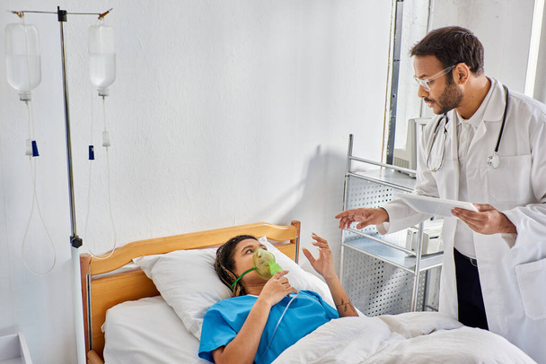 chora Afroamerykanka leżąca w łóżku w masce tlenowej patrząc na indyjskiego lekarza trzymającego tabletkę - Zdjęcie, obraz