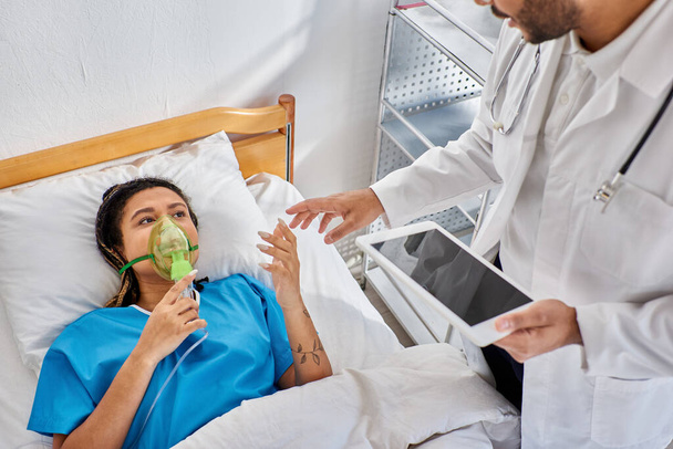 mujer afroamericana enferma acostada en la cama con máscara de oxígeno mirando a su médico indio sosteniendo la tableta - Foto, Imagen