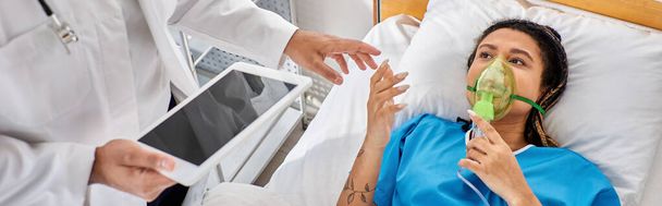 Afrikaans-Amerikaanse vrouw liggend in bed met zuurstofmasker kijkend naar Indiase arts met tablet, spandoek - Foto, afbeelding