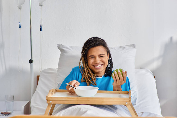 νεαρή Αφροαμερικανή που τρώει χυλό και μήλο για πρωινό στο νοσοκομείο, υγειονομική περίθαλψη. - Φωτογραφία, εικόνα