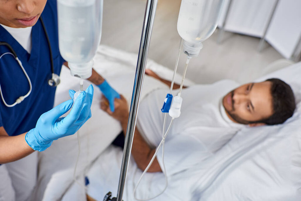 concentrarsi su infermiera afro-americana impostazione drop counter accanto a uomo offuscato sdraiato nel letto d'ospedale - Foto, immagini