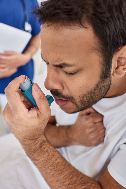 bijgesneden zicht op indische man met astma-inhalator met wazig verpleegkundige op de achtergrond, gezondheidszorg - Foto, afbeelding