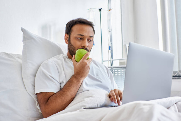 joven indio hombre teniendo videollamada y sosteniendo manzana verde mientras está acostado en la cama del hospital, atención médica - Foto, Imagen
