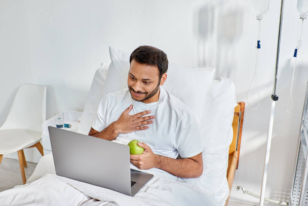 молодой индиец, имеющий видео-звонок и держащий зеленое яблоко во время лежания в больничной койке, здравоохранения - Фото, изображение