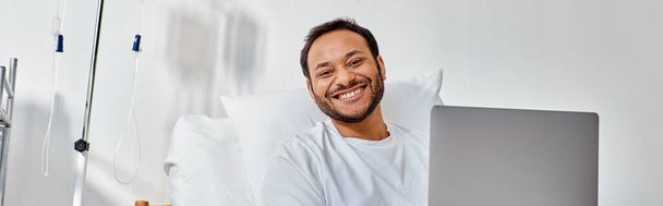 Genç, neşeli Hintli adam hastane yatağında dizüstü bilgisayarla çalışırken kameraya gülümsüyor. - Fotoğraf, Görsel