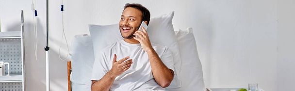 alegre joven indio hombre hablando alegremente por teléfono mientras está acostado en la cama en la sala de hospital, pancarta - Foto, Imagen