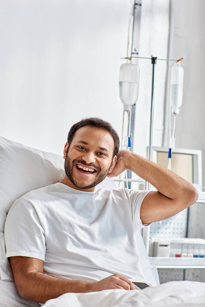 alegre joven indio acostado en la cama del hospital y sonriendo alegremente a la cámara, cuidado de la salud - Foto, Imagen