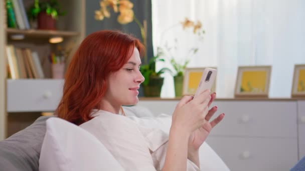 若い美しい女性はスマートフォンを使用し,ベッドに横たわっている間に友人とニュースやテキストを読みます - 映像、動画