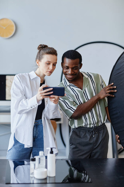 Retrato vertical de dois jovens fotógrafos trabalhando em fotografia de produto em estúdio e usando telefone celular para mídias sociais - Foto, Imagem