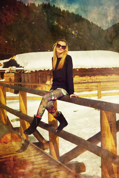 Beautiful young blonde lady posing on a wooden balustrade in a snowy winter landscape. - Photo, Image