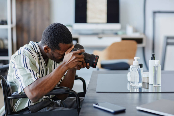 Widok z boku portret młodego czarnego człowieka na wózku inwalidzkim jako fotograf z niepełnosprawnością biorąc zdjęcia produktów w studio fotograficznym - Zdjęcie, obraz