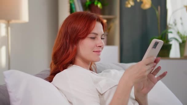mujer sonriente se comunica con amigos usando mensajeros instantáneos en el teléfono móvil mientras se sienta en el sofá en la habitación - Metraje, vídeo