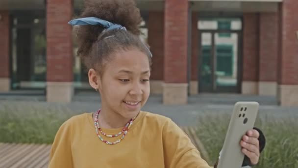 Klein Afro-Amerikaans meisje in beschermende pols bewakers met mobiele telefoon, glimlachen en chatten op video bellen terwijl ze buiten rust na rolschaatsen - Video