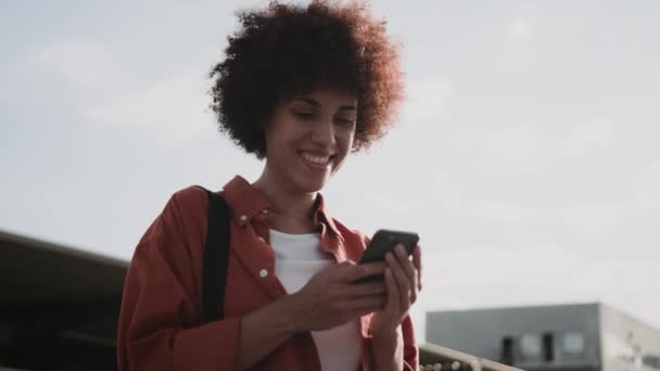 Счастливая молодая афроамериканка, печатающая по мобильному телефону, ожидая такси в городе. Люди, образ жизни, концепция путешествий. Медленное движение - Кадры, видео