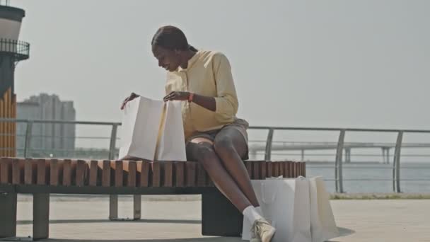 Plan complet de femme afro-américaine joyeuse assise sur un banc à l'extérieur sur le bord de la rivière de la ville, regardant dans des sacs à provisions et souriant - Séquence, vidéo