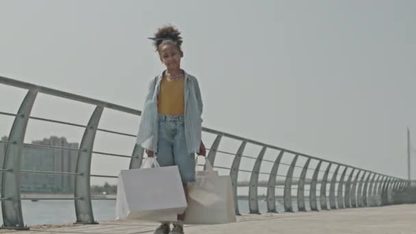 Ampia ripresa della piccola ragazza afroamericana in piedi sul lungofiume della città con borse della spesa e in posa per la fotocamera durante la soleggiata giornata estiva - Filmati, video