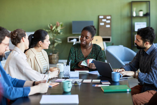 Zelfverzekerde Afro-Amerikaanse zakenvrouw met palet die haar mening uitlegt aan collega 's terwijl ze naar een van hen kijkt tijdens een ontmoeting - Foto, afbeelding