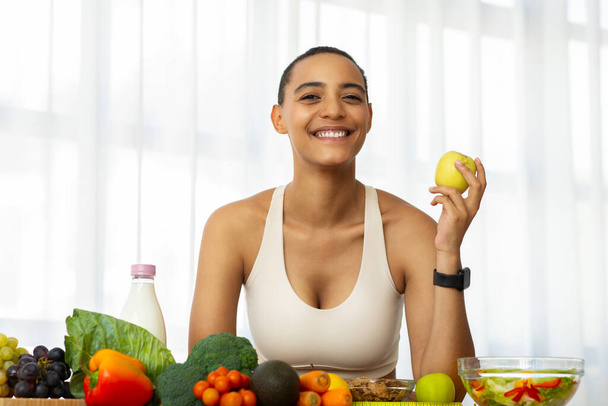 Spor giyim kuşağındaki neşeli Latin bayan açık mutfak içlerinde yeşil elma yiyor, taze sebzelerin ve salatanın tadını çıkarıyor. Diyet, kilo kaybı, sağlıklı yiyecekler, vejetaryen ve vitaminler - Fotoğraf, Görsel