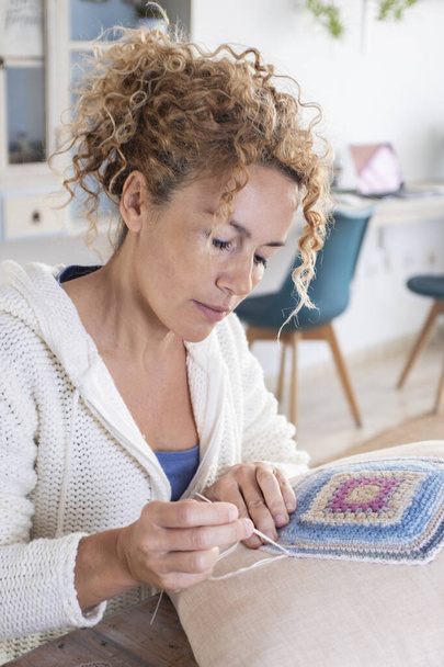 Adult caucasian woman at home doing crochet hobby activity at home decorating a shabby chic white pillow. Living room interior in background. Female people creating woolen decorations - Fotó, kép