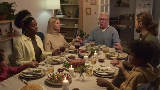Familia multiétnica tomándose de las manos y rezando mientras están sentados en la mesa festiva durante la celebración de Acción de Gracias en casa - Imágenes, Vídeo