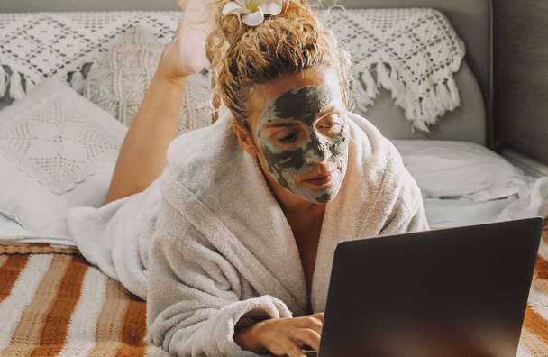 Pretty adult woman with skin beauty cream on face work on laptop computer laying in camper van bedroom. Travel lifestyle female people with online job. Modern lady and healthy treatment - Foto, Imagen