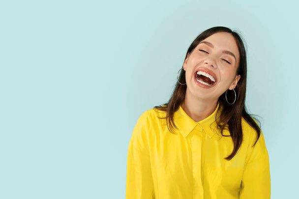 Fröhlich funky attraktive junge brünette Frau in gelbem Hemd lachen isoliert auf blauem Studiohintergrund, posiert neben leeren Kopierraum. Konzept menschlicher Emotionen - Foto, Bild