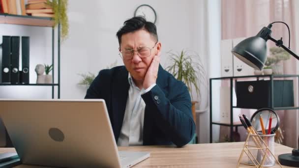 Zahnprobleme. Asiatischer Geschäftsmann, der im Büro arbeitet, berührt die Wange, schließt die Augen mit dem Ausdruck der schrecklichen leiden unter schmerzhaften Zahnschmerzen, empfindlichen Zähnen, Hohlräumen. Männliche Chinesen im formellen Anzug - Filmmaterial, Video