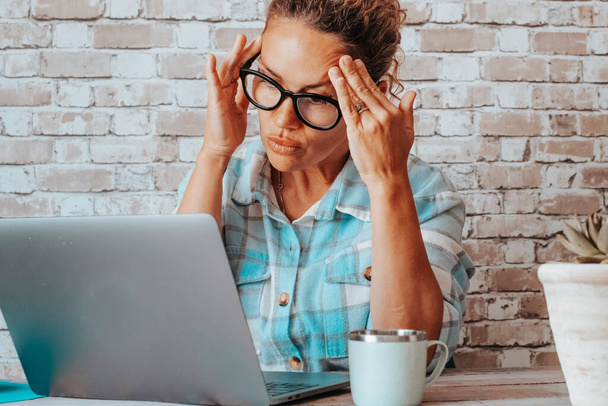 Egy nő, aki a laptopnál dolgozik fejfájással. Koncentrált és aggódó női üzletasszony egy számítógép előtt. Házi munka fali téglákkal a háttérben. Online állás- és biztonsági koncepció - Fotó, kép