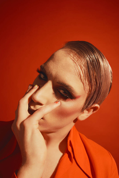 Makeup artist. Portrait of handsome young man, gay with bright stylish makeup, hairstyle posing on red studio background. Concept of male makeup, fashion, lgbtq community, self-identity, acceptance - 写真・画像