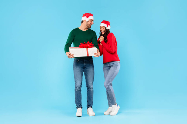 Kerstverrassing. Man geeft kerstcadeau doos aan vrouw vieren Nieuwjaar, het dragen van Santa hoeden, staande over blauwe studio achtergrond. Vakantie verkoop en aanbiedingen. Volledige lengte schot - Foto, afbeelding
