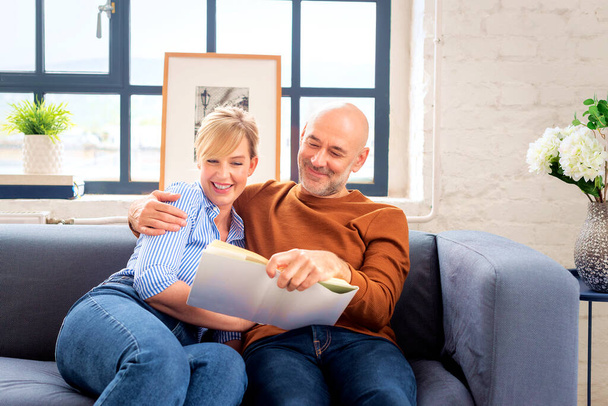 Shot of a married couple reading a book together and relaxing on the sofa at home. Blond haired woman and mid aged man wearing casual clothes.  - Photo, image