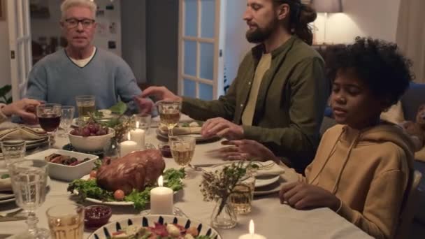 Interkulturní členové rodiny se drží za ruce a modlí se před slavnostní večeří v útulném obývacím pokoji - Záběry, video