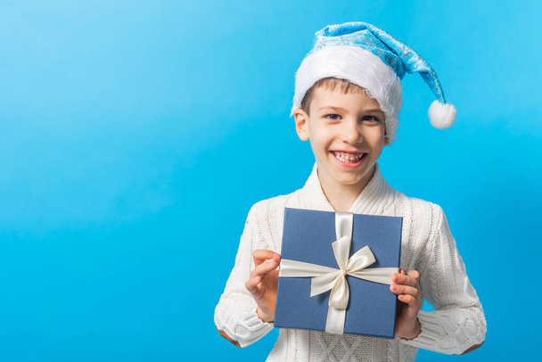 Happy little boy in blue Christmas hat with a gift box copy space. Empty place for your text. Winter holidays and present concept - Photo, Image