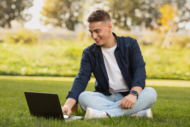 Радостный кавказский мужчина тысячелетия наслаждается образом жизни и отдыхом, печатая на ноутбуке, расслабляясь в парке, садясь на траву. Работа, бизнес, учеба с технологиями, фриланс на свежем воздухе, перерыв в свободное время, цифровая реклама - Фото, изображение