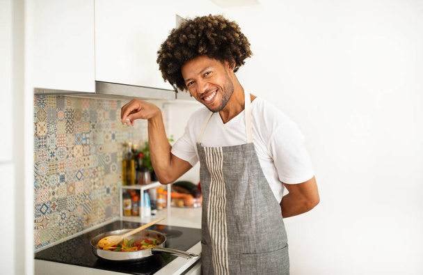 Ragazzo afroamericano stagioni uno stufato di verdure sui fornelli, sorridendo alla fotocamera in cucina moderna, incarnando una sana alimentazione. L'uomo padroneggia la cucina vegetariana, cospargendo spezie per il sapore - Foto, immagini