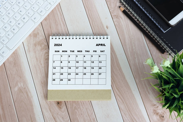 Biały kwiecień 2024 kalendarz na drewnianym biurku z klawiaturą, notatnikami, ołówkiem, rośliną doniczkową i smartfonem. - Zdjęcie, obraz