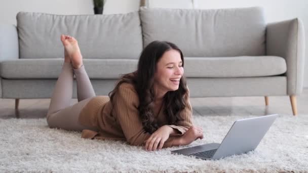 Caucasian woman lies on the living room floor, her laughter filling the room as she engages with laptop. Eyes sparkle with happiness, creating an atmosphere of warmth and mirth. - Footage, Video