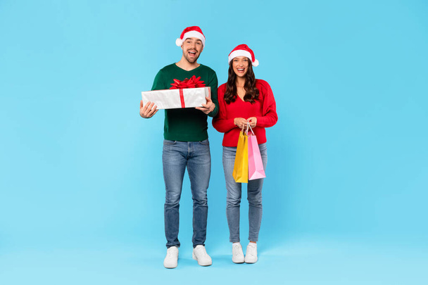 Cadeaux de Noël Shopping. Happy Couple tenant des sacs Shopper et enveloppé boîte cadeau debout sur fond bleu studio, souriant à la caméra. Offres de vente de vacances. Longueur totale - Photo, image